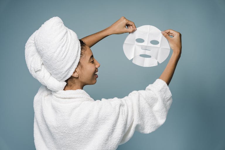 Fresh Content Daily Back view of happy young African American female in bathrobe with towel on head smiling while demonstrating skin care mask against blue background