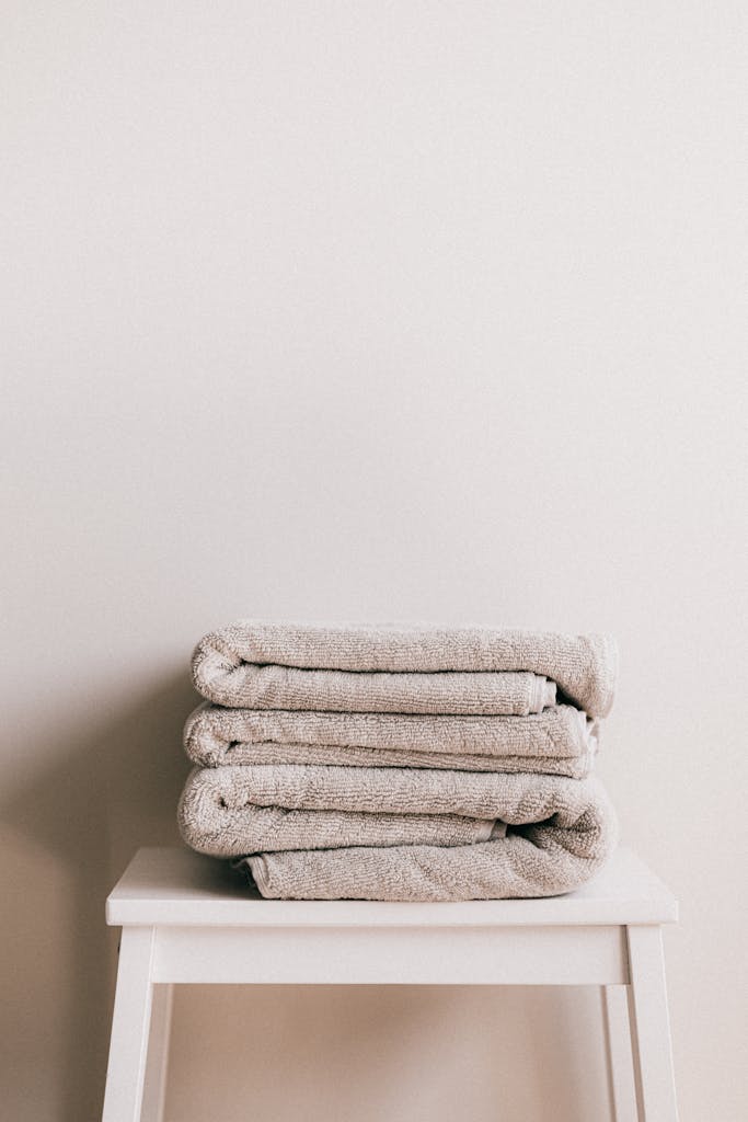 Daily Blog composition of white simple stool with clean fresh folded gray towels against beige wall in bathroom