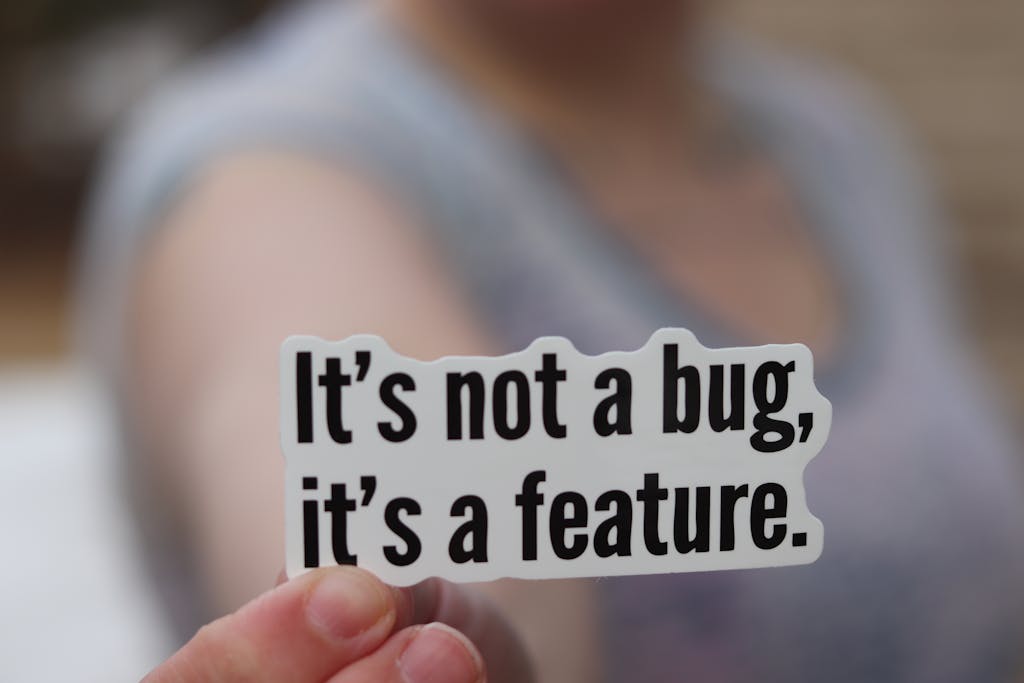 Sydneebeeyxo Close-up of a hand holding a sticker with a humorous tech message: 'It's not a bug, it's a feature.'