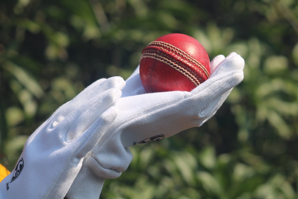 Wheon Cricket 07 Detailed photo of a red cricket ball held by gloved hands outdoors.