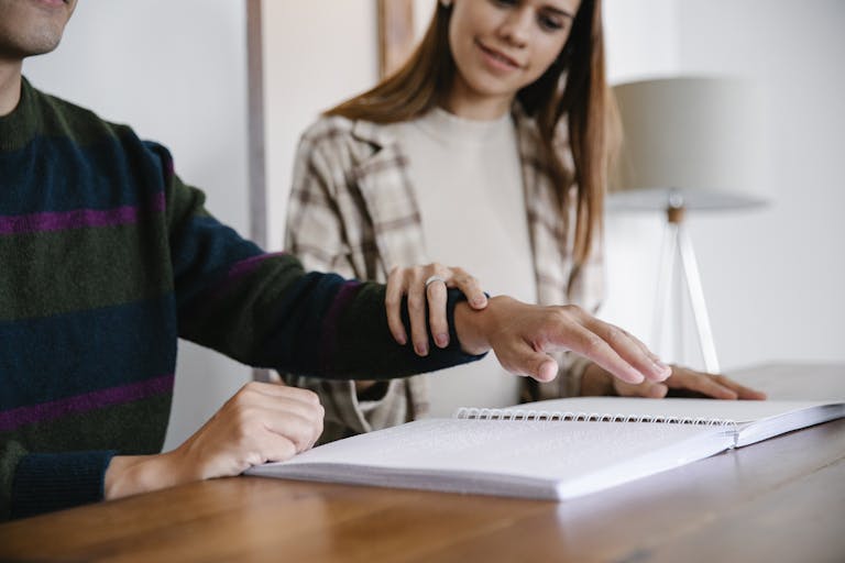 Dainikshiksha.com Person guiding another's hand over a Braille book, showing support and assistance indoors.
