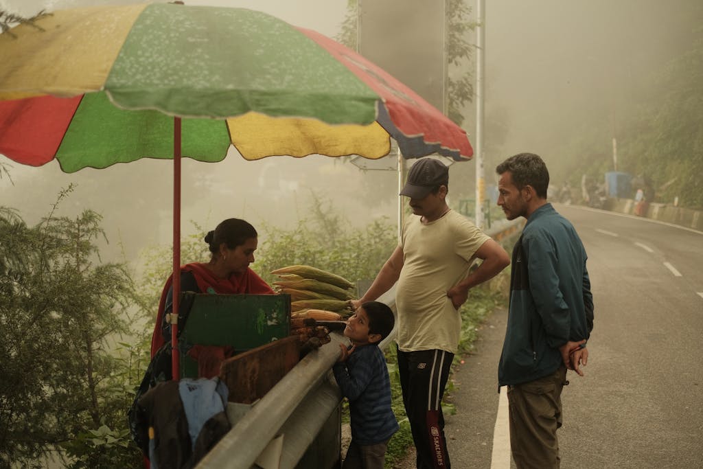 R6 Market Place A family buys corn from a vendor under colorful umbrella on a misty rural road.