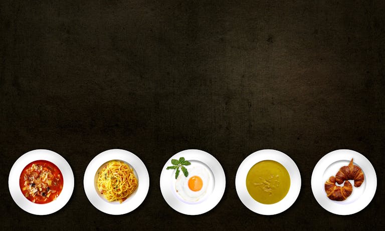 sodiceram Flatlay of various dishes including pasta, egg, soup, and croissant on textured background.