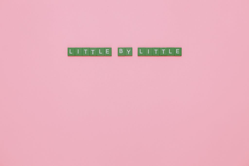 ventsMagazine Green tiles spelling 'LITTLE BY LITTLE' on a pink background conveying motivation.